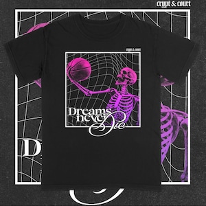 以下が含まれることがあります： 黒いTシャツ。ネオンピンクの骸骨がバスケットボールを白いフレーム内で持っています。「Dreams Never Die」の文字が下に表示されています。「crypt & court」のブランド名が上部に印刷されています。