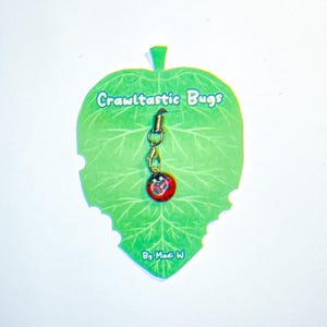 May include: A red and black ladybug charm with a gold chain and clasp. The charm is attached to a green leaf-shaped card with the text "Crawltastic Bugs" and "By Madi W".