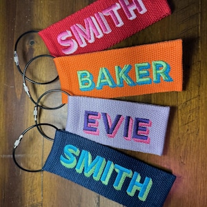 K&ouml;nnte beinhalten: Vier personalisierte Gep&auml;ckanh&auml;nger in Rot, Orange, Lavendel und Marineblau. Jeder Anh&auml;nger zeigt einen anderen Namen, der in kontrastierenden Farben gestickt ist: SMITH, BAKER, EVIE und SMITH. Die Anh&auml;nger sind an Metall-Schl&uuml;sselringen befestigt.
