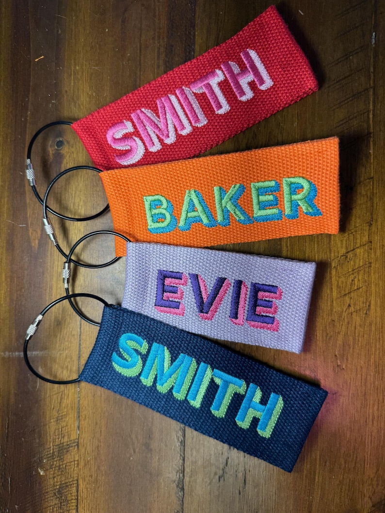 K&ouml;nnte beinhalten: Vier personalisierte Gep&auml;ckanh&auml;nger in Rot, Orange, Lavendel und Marineblau. Jeder Anh&auml;nger zeigt einen anderen Namen, der in kontrastierenden Farben gestickt ist: SMITH, BAKER, EVIE und SMITH. Die Anh&auml;nger sind an Metall-Schl&uuml;sselringen befestigt.