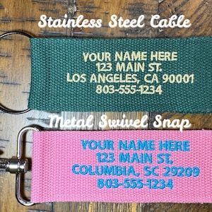 K&ouml;nnte beinhalten: Zwei personalisierte Schl&uuml;sselanh&auml;nger. Einer ist gr&uuml;n mit einem Edelstahlkabel und goldenem Text. Der andere ist rosa mit einem Metall-Karabinerhaken und blauem Text. Beide haben den Text "YOUR NAME HERE", Adresse und Telefonnummer.