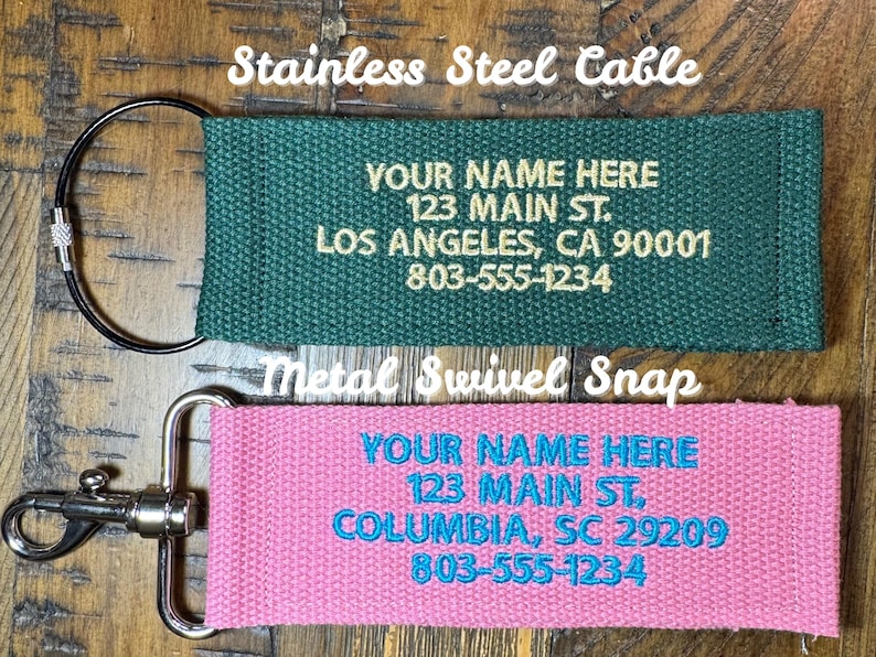 K&ouml;nnte beinhalten: Zwei personalisierte Schl&uuml;sselanh&auml;nger. Einer ist gr&uuml;n mit einem Edelstahlkabel und goldenem Text. Der andere ist rosa mit einem Metall-Karabinerhaken und blauem Text. Beide haben den Text "YOUR NAME HERE", Adresse und Telefonnummer.