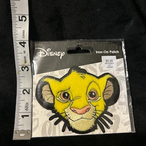 May include: A yellow and black embroidered iron-on patch featuring the face of Simba from The Lion King, positioned next to a measuring tape showing the patch is approximately 3.5 inches tall. The packaging is labeled Disney Iron-On Patch.