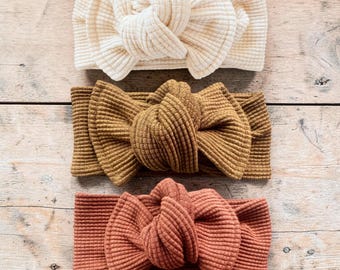 May include: Three ribbed headbands arranged vertically. The top headband is cream, the middle is mustard yellow, and the bottom is terracotta orange. Each headband features a central knotted bow detail made from the same ribbed fabric.