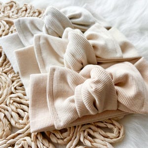 May include: Three beige ribbed fabric hair bows arranged in a stack. The bows are made of a soft, stretchy material and are perfect for adding a touch of style to any outfit.