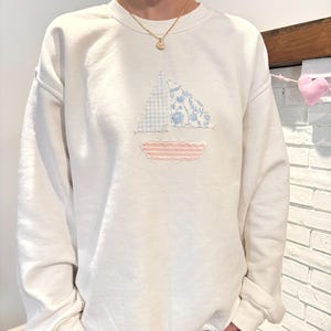 May include: White sweatshirt with a patchwork sailboat design in blue and pink on the front.