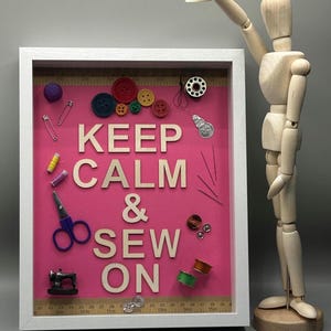 Puede incluir: Una caja de sombra blanca con un fondo rosa y el texto "KEEP CALM & SEW ON". La caja está llena de varios artículos de costura, incluidos botones, agujas, hilo y una pequeña máquina de coser.