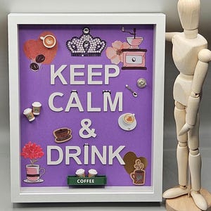 May include: A white framed picture with a purple background and the text "Keep Calm & Drink" in white letters. The picture is decorated with coffee cups, coffee beans, a crown, a flower, and a coffee grinder.