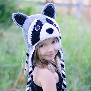 May include: A child wearing a gray, white, and black crocheted raccoon hat with black and white braided ties. The hat has a black nose, white fur, and black eyes.