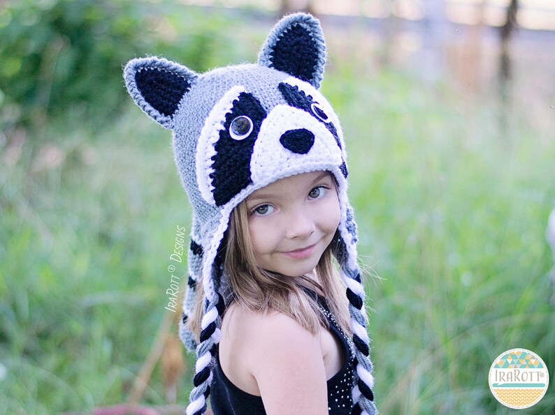 May include: A child wearing a gray, white, and black crocheted raccoon hat with black and white braided ties. The hat has a black nose, white fur, and black eyes.