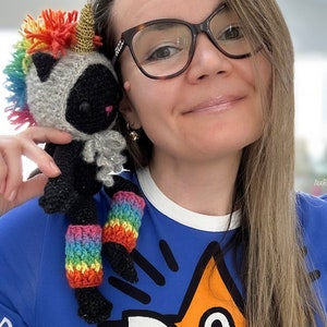 May include: A woman wearing a blue shirt with a cartoon fox design is holding a crocheted black cat wearing a rainbow unicorn horn and leg warmers. The cat has black eyes and a white face.