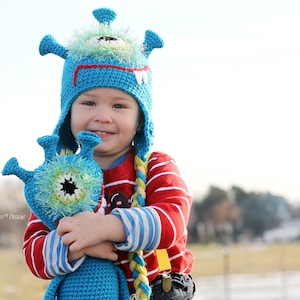 Peut inclure: Un jeune enfant portant un chapeau de monstre en crochet bleu et vert avec un jouet de monstre en crochet assorti. Le chapeau a une longue queue tressée.
