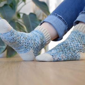 May include: A pair of handmade crochet socks in shades of blue, beige, and white. The socks feature an openwork design and are worn with blue jeans. The toe and heel are solid beige, while the body has a textured, open weave.