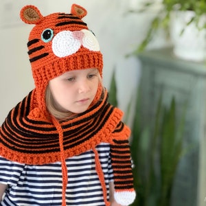 May include: A child wearing a crocheted orange and black tiger hat and scarf. The hat has a white face with blue eyes and a pink nose.