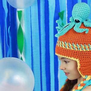 May include: A child wearing a crocheted orange and yellow hat with a turquoise octopus on top. The hat has ear flaps and a braided chin strap. The child is looking at a clear balloon.