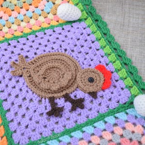 Pode incluir: Uma manta de croch&ecirc; com um quadrado roxo que apresenta um frango de croch&ecirc; marrom com uma crista vermelha. A manta &eacute; feita com uma variedade de cores e texturas, incluindo verde, rosa e amarelo.