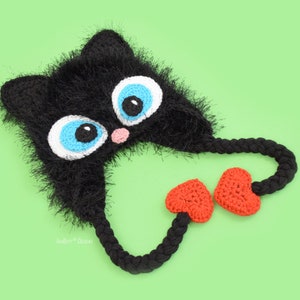 May include: A handmade black crochet cat hat with large blue and white eyes, a pink nose, and orange heart-shaped accents. The hat has long braided ties and is set against a light green background. The hat is a whimsical accessory.