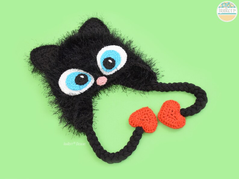 May include: A handmade black crochet cat hat with large blue and white eyes, a pink nose, and orange heart-shaped accents. The hat has long braided ties and is set against a light green background. The hat is a whimsical accessory.