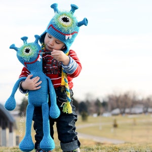 May include: A young child wearing a blue crocheted monster hat holds a blue crocheted monster toy. The hat and toy have large, green eyes and white teeth.