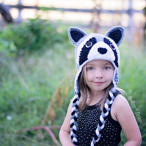 May include: A child wearing a gray and white crocheted raccoon hat with black and white braided ties. The hat has black eyes, a black nose, and white teeth.