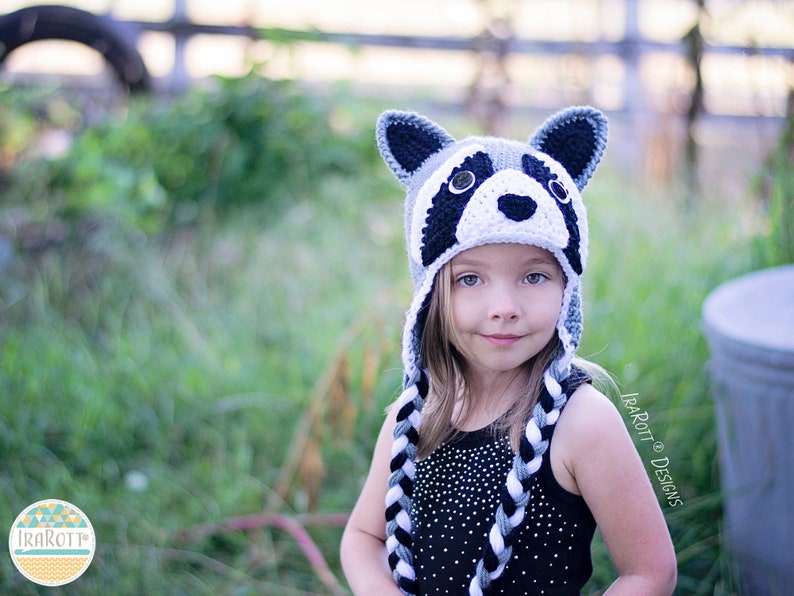 May include: A child wearing a gray and white crocheted raccoon hat with black and white braided ties. The hat has black eyes, a black nose, and white teeth.