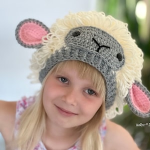 May include: A child wearing a gray and white crocheted sheep hat with pink ears. The hat has a fluffy white wool texture and a gray face with black eyes.