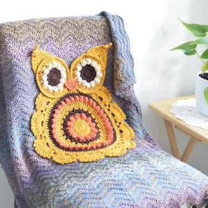 May include: A handmade crochet owl pillow in shades of yellow, orange, and brown rests on a multi-colored blanket. The blanket is draped over a chair, with a plant in a white pot on a small wooden table in the background.