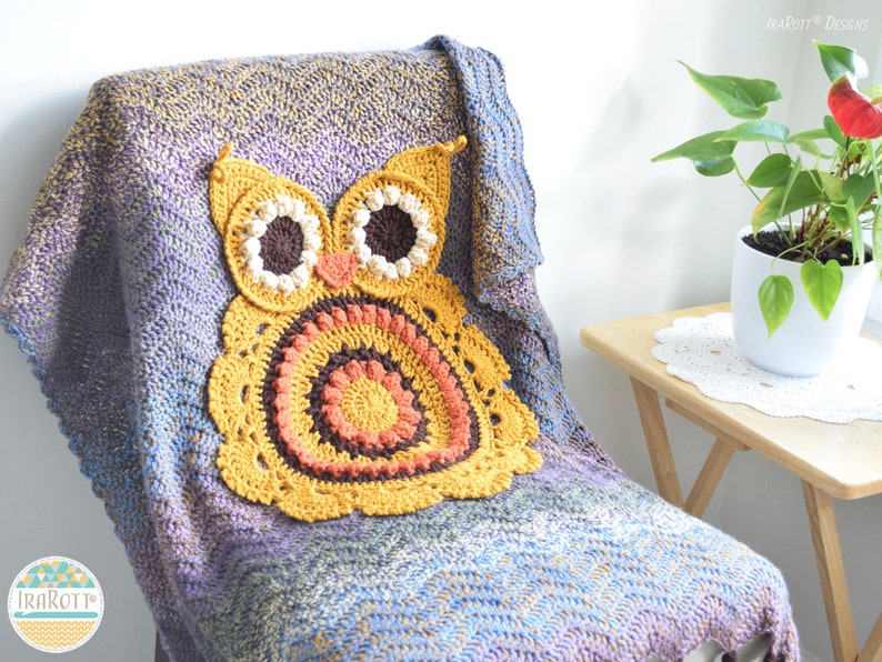 May include: A handmade crochet owl pillow in shades of yellow, orange, and brown rests on a multi-colored blanket. The blanket is draped over a chair, with a plant in a white pot on a small wooden table in the background.
