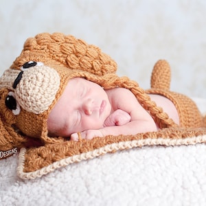 May include: A newborn baby wearing a crocheted brown and beige dog costume, sleeping on a white blanket. The costume features a dog's head with black eyes and a brown nose.