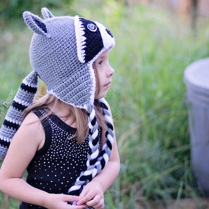 May include: A child wearing a gray and black crocheted raccoon hat with a long tail. The hat has a white stripe around the eyes and a black button nose.