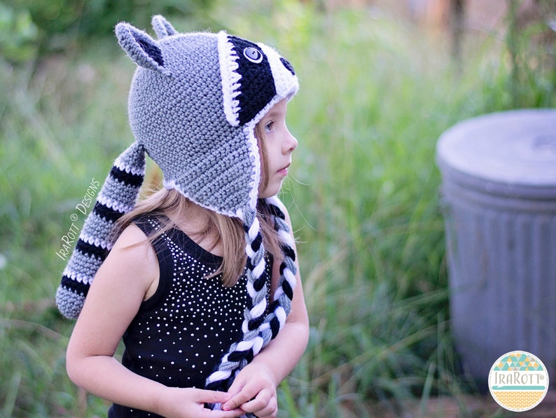 May include: A child wearing a gray and black crocheted raccoon hat with a long tail. The hat has a white stripe around the eyes and a black button nose.