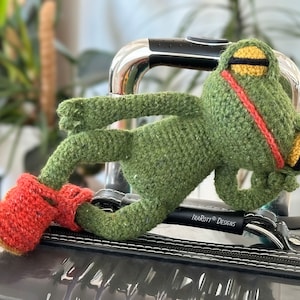 May include: A green crocheted frog toy with yellow and black accents is resting on a black suitcase with the text "iBAROTT Designs". The frog is wearing red crocheted boots.