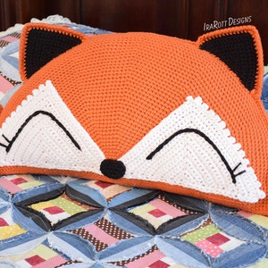 May include: A crocheted fox-shaped cushion with orange fur, white eyes, and black nose and ears. The cushion is lying on a colourful patchwork quilt.