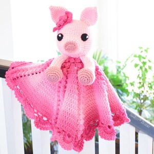 Puede incluir: Una manta de bebé de crochet rosa con un cerdo de crochet rosa adjunto. La manta tiene un borde festoneado y el cerdo tiene una flor rosa en la cabeza.