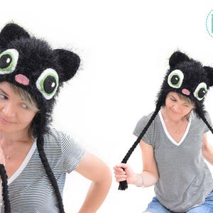 May include: A black cat hat with large, cartoonish eyes, ears, and a pink nose. The hat has long, braided ties. The hat is worn by a person wearing a striped shirt.