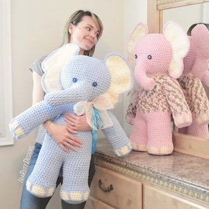 May include: Two crocheted elephant plushies, one blue and one pink, with white ears and blue eyes. The blue elephant is being held by a person.