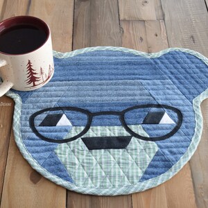 May include: A blue denim and green plaid quilted coaster shaped like a bear's face with black glasses.