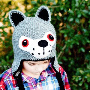 May include: A gray crocheted wolf hat with black ears and a white face. The hat has red eyes and a black nose. A child is wearing the hat.