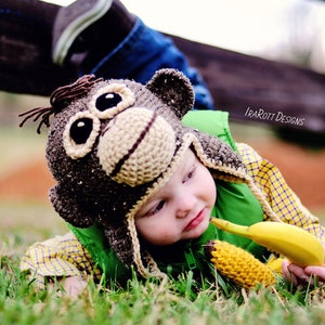 May include: A young child wearing a brown and beige monkey hat with a green vest and yellow plaid shirt is lying in the grass. The child is holding a yellow banana and a yellow crocheted banana.