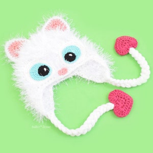 May include: A handmade white crochet cat hat with pink accents on the ears and nose, and large blue eyes with black pupils. The hat has long white braided ties ending in pink heart-shaped embellishments. The hat is on a light green background.