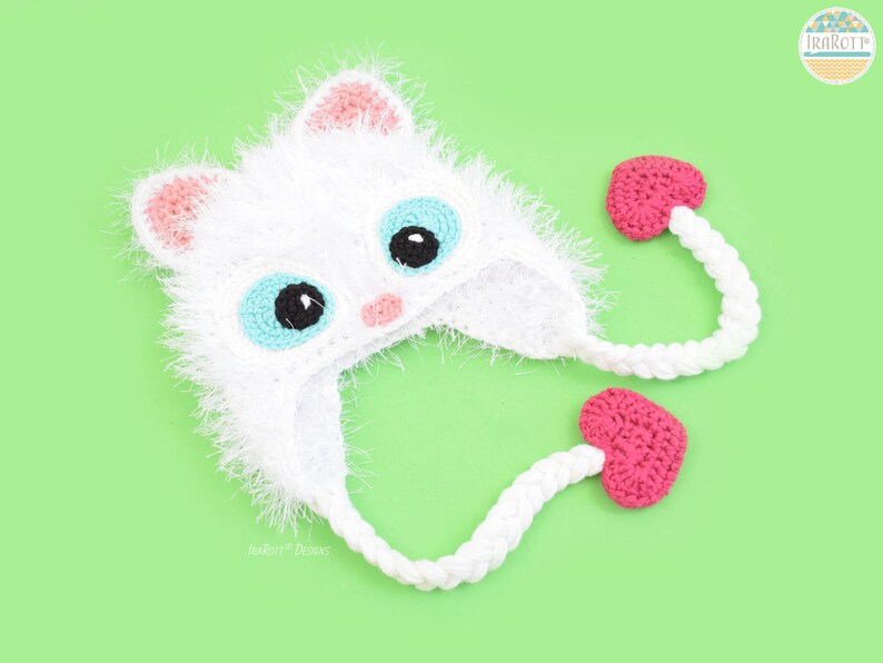May include: A handmade white crochet cat hat with pink accents on the ears and nose, and large blue eyes with black pupils. The hat has long white braided ties ending in pink heart-shaped embellishments. The hat is on a light green background.