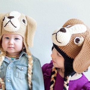 May include: Two children wearing crocheted dog hats with long braided tails. The hats are brown and beige with white and black details. The children are looking at each other.