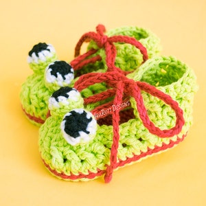 May include: A pair of green crocheted baby booties with red laces and frog-like eyes. The booties have a red trim around the edges.