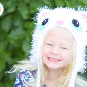 May include: A white crocheted cat hat with black, blue, and pink details. The hat has long white braids and a fluffy trim. The image includes the text "IvaRott Designs".