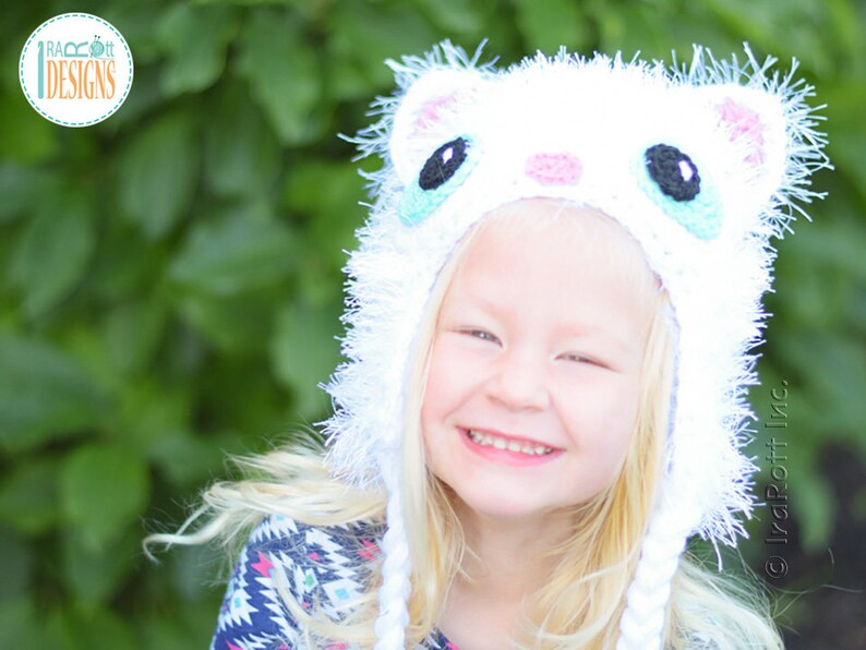 May include: A white crocheted cat hat with black, blue, and pink details. The hat has long white braids and a fluffy trim. The image includes the text "IvaRott Designs".