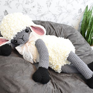 Op de afbeelding: Een groot gebreid grijs en wit schaap knuffeldier met zwarte hoeven en roze oren. Het schaap ligt op een grijze zitzak met een witte, bloemige achtergrond.