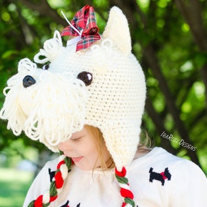May include: A white crocheted dog hat with a red and green plaid bow on top. The hat has a long white yarn beard and floppy ears. The hat is being worn by a child wearing a white sweater with a black and white dog design.