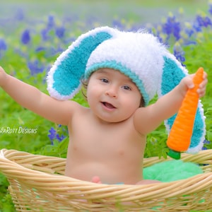 May include: A baby wearing a white and turquoise bunny hat holds a toy carrot while sitting in a wicker basket. The baby is surrounded by bluebonnet flowers.