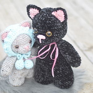 May include: Two crocheted cat plushies, one gray with a blue hat and the other black with a pink ribbon.
