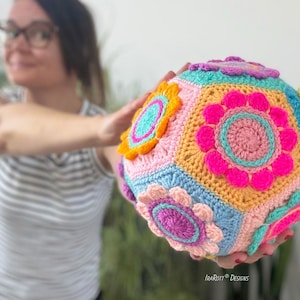 May include: A colorful, handmade crochet ball featuring a geometric design with floral accents. The ball is composed of various pastel colors, including pink, blue, yellow, and orange. The ball is approximately 6 inches in diameter.